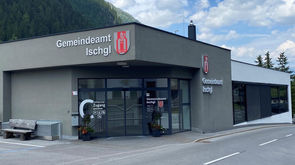 The image shows the municipality office of Ischgl, a modern building with large windows. In the background, mountains and a blue sky can be seen.