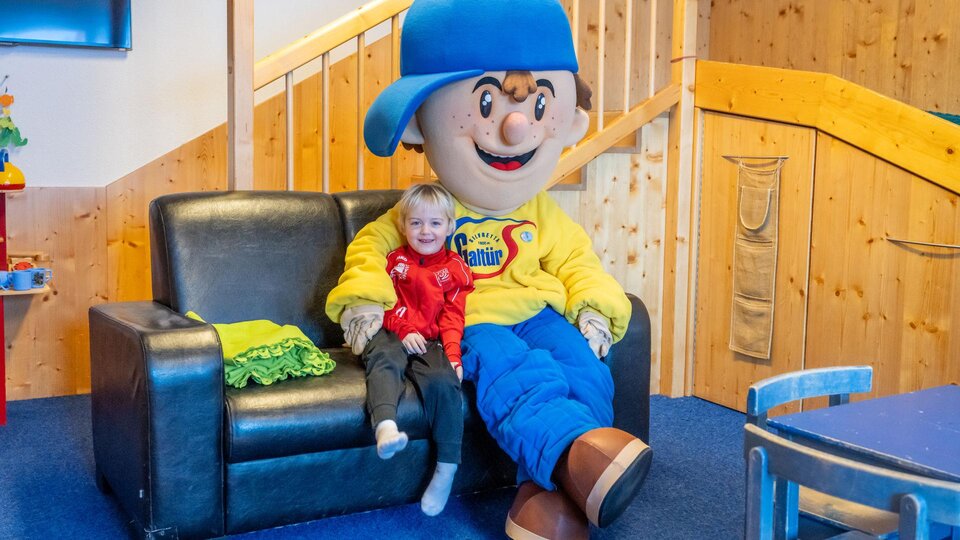 A child is sitting smiling next to a large figure with a blue hat. The scene takes place in a cozy playroom.