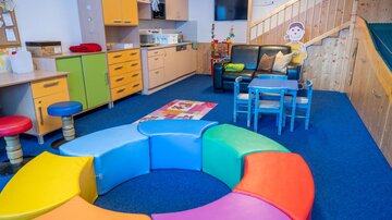 A colorful play area with a colorful seating arrangement in the shape of a circle. In the background, there is a kitchen and a cozy corner for children.