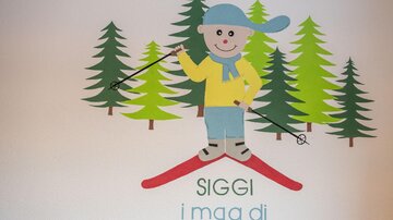 A cheerful skier with a blue hat and yellow sweater stands on red skis. In the background, green trees can be seen.