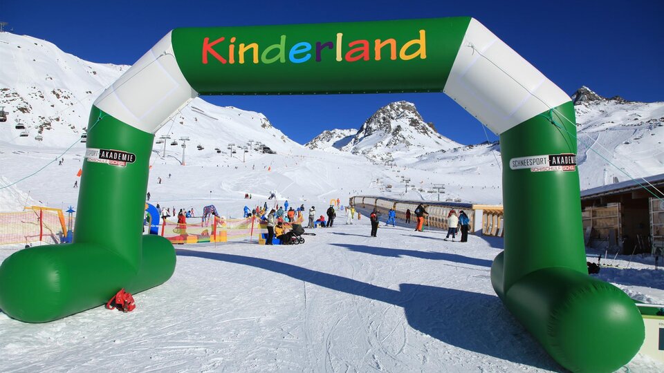 A colorful arc with the inscription "Kinderland" stands in a snowy ski resort. In the background, ski lifts and skiers can be seen.