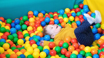 A small child is happily lying in a ball pit. They are surrounded by colorful balls in various colors.