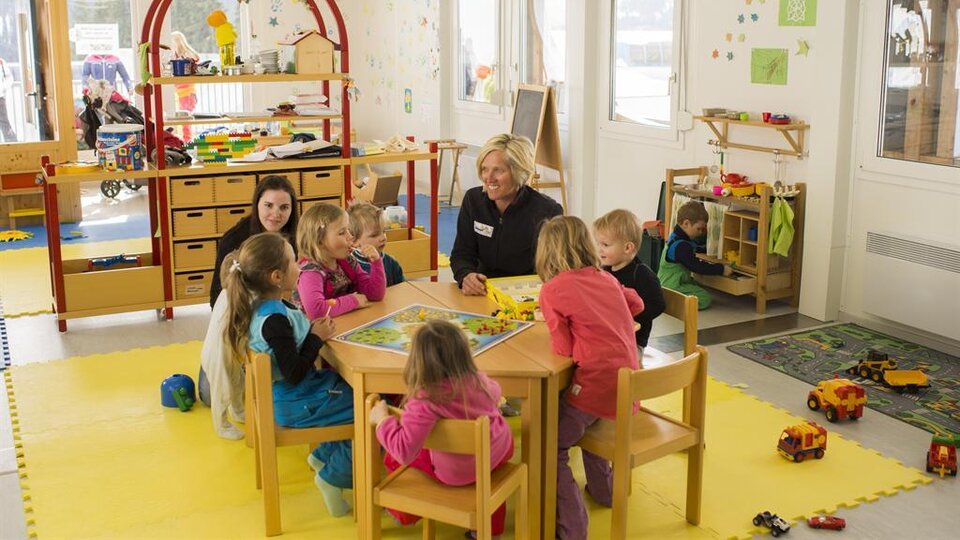 A bright kindergarten room with children sitting around a table and playing. A caregiver is present, while in the background, other children are occupied with toys.