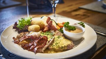 A plate with hearty food, including meat, bacon, and vegetables. Next to it, there is a tasty sauce and potatoes.