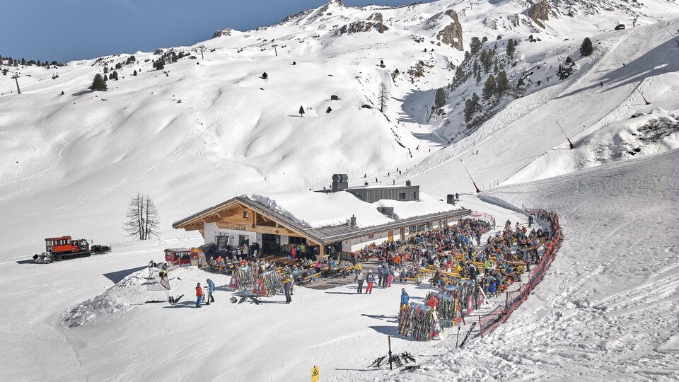 A bustling ski resort with a large restaurant amidst snow-covered mountains. Many people are enjoying the sun and winter activities.