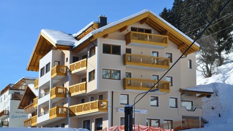 Ein modernes Hotelgebäude mit Holzbalkonen in einer schneebedeckten Umgebung. Im Hintergrund sind Tannenbäume und ein klarer blauer Himmel zu sehen.