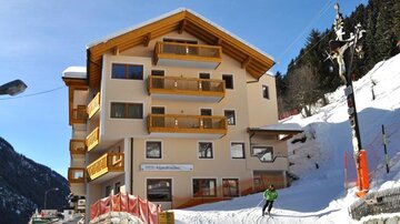 Ein gemütliches Hotel in den Bergen mit einem schneebedeckten Hang. Im Hintergrund sind Bäume und ein klarer Himmel zu sehen.
