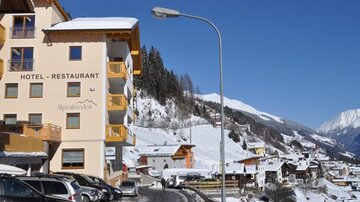 Ein Hotel in einer verschneiten Berglandschaft, umgeben von schneebedeckten Bergen und kleinen Häusern. Die Straße vor dem Hotel ist schmal und gut befahrbar.