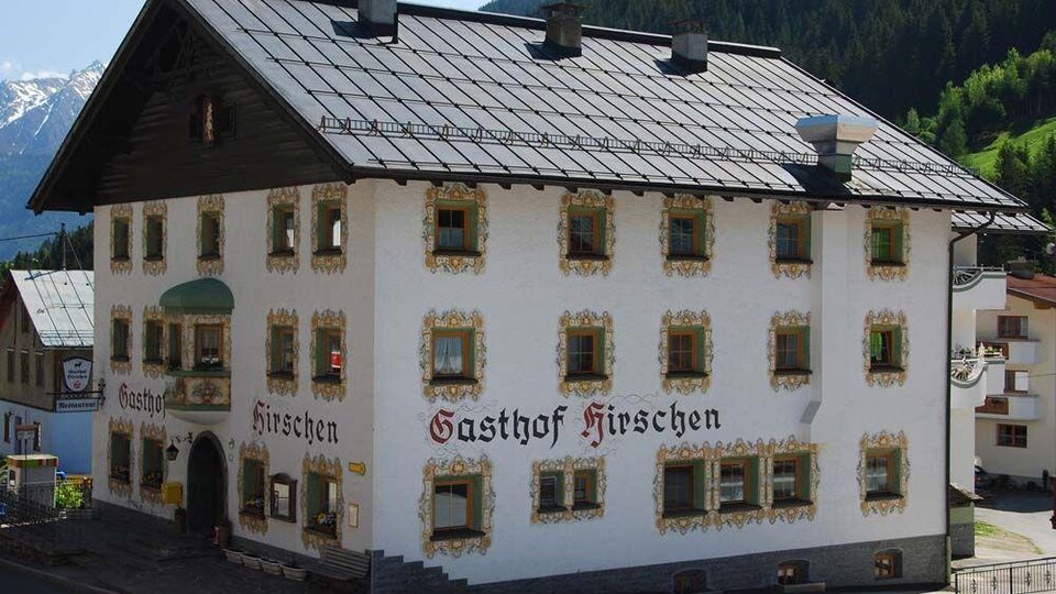 A traditional inn with the sign "Gasthof Hirschen" on the facade. The building has historical architecture and is located in a picturesque mountain landscape.