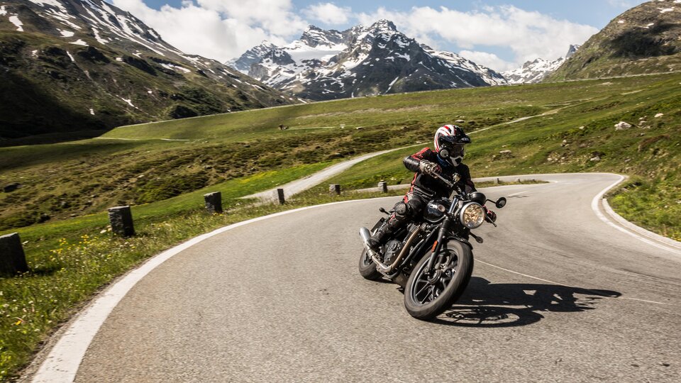 Ein Motorradfahrer fährt auf einer kurvigen Straße in den Bergen. Die Landschaft ist grün mit schneebedeckten Gipfeln im Hintergrund.