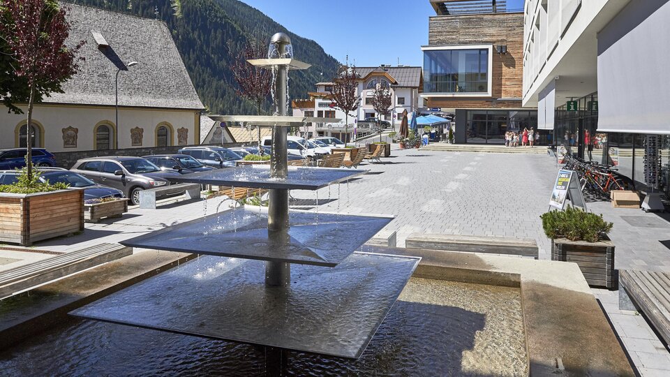 A modern square with a water basin and several levels. In the background, cars and buildings can be seen, surrounded by mountains.