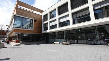 A modern building with large windows and wood cladding. The square in front is paved and offers seating options.