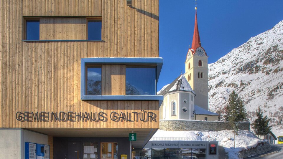 A modern community house with a wooden facade in Galtür. In the background, a church with a red bell tower and snow-covered mountains can be seen.