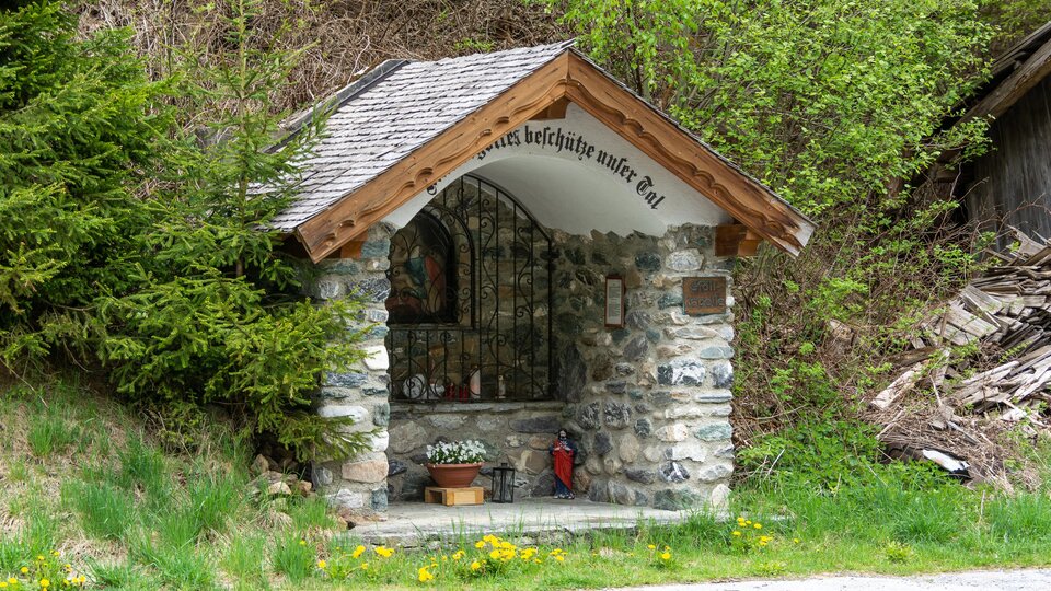 A small stone chapel with a wooden roof stands in nature. Surrounded by trees and grass, it offers a peaceful retreat.