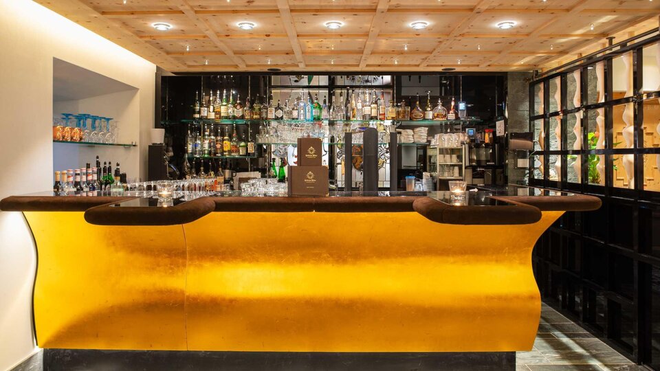 An elegant bar with a golden counter and a variety of spirits. The wall shelves are decorated with glasses and drinks.