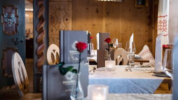 A cozy restaurant with wooden interiors and elegantly set tables. On the tablecloths are menus, candles, and red roses.