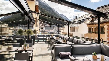An elegant rooftop terrace with comfortable seating and tables. The view includes beautiful mountains and a charming town.