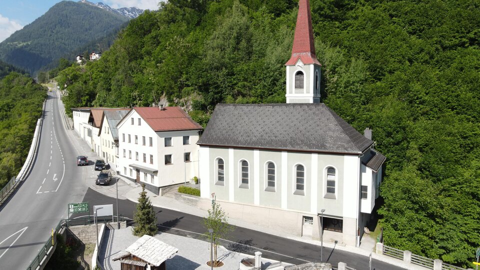 A small church with a red roof and a tall tower stands by a road. Surrounded by green hills and residential buildings.