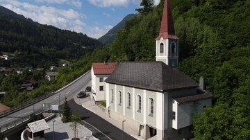 A picturesque church with a tall red tower, surrounded by green hills. The road runs nearby and there are some trees and buildings in the vicinity.