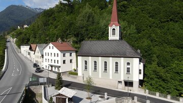 A small church with a red tower stands beside a road, surrounded by green trees. In the background, mountains can be seen, and the architecture is typical for a rural area.