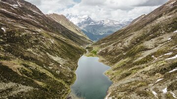 A picturesque mountain landscape with a clear lake between high mountains. Clouds are over the valley, and the snow-capped peaks are visible in the distance.