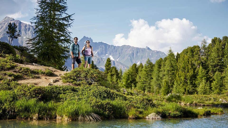 Two hikers stand at the shore of a clear lake, surrounded by green meadows and tall mountains. The sky is blue with some clouds.