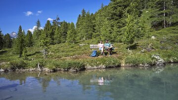 A peaceful landscape with clear blue water and green trees. Two people are sitting relaxed on a bench by the shore.