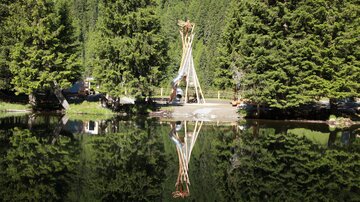 A beautiful pond surrounded by tall, green trees. In the foreground, a wooden sculpture can be seen, reflecting in the water.