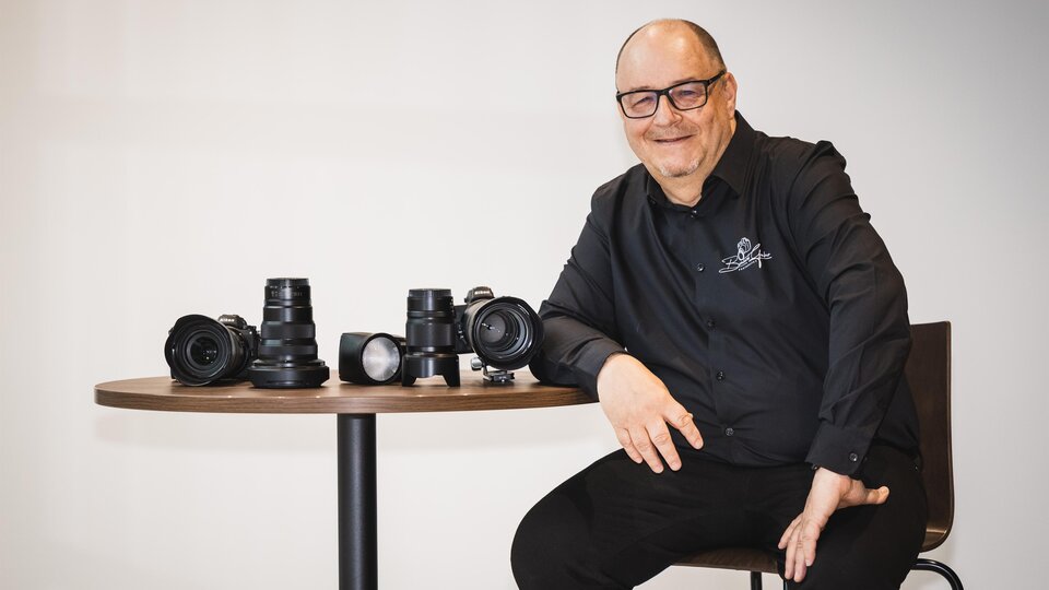 A smiling man sits at a table and presents several cameras and lenses. He is wearing a black shirt and appears friendly and competent. | © Bernhard Gruber – Photography