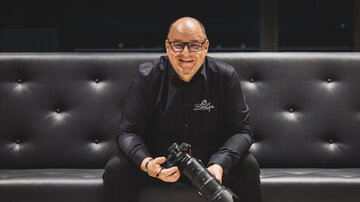 A smiling man is sitting on a dark couch and holding a camera in his hand. He is wearing a black shirt and appears friendly and approachable. | © Bernhard Gruber – Photography