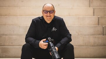 A photographer sits on a staircase holding a camera body in his hand. He is wearing a black shirt and smiling at the camera. | © Bernhard Gruber – Photography
