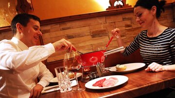 A couple enjoys a fondue dinner in a cozy restaurant. On the table are various ingredients and glasses of wine.
