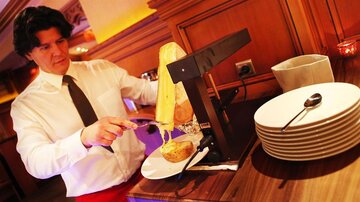 A waiter is serving melted cheese from a cheese block. In the background, empty plates and a bowl are visible.