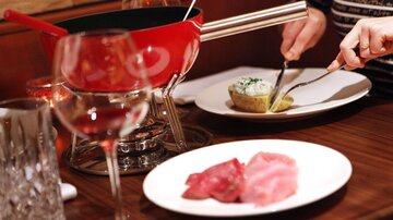 A table with a red fondue pot and various types of meat on a white plate. In the background, there is a glass of wine and a serving of potatoes with herb quark.
