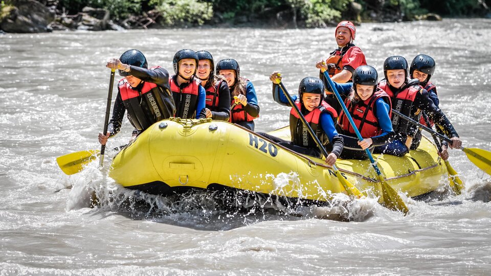 A group of eight people is paddling in a yellow raft on a river. They are wearing helmets and life jackets and having a lot of fun.