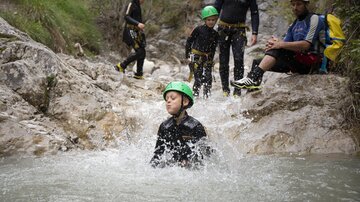 A group of children and adults is having fun with water sports in a river. Some are wearing green safety helmets and playing in the water.