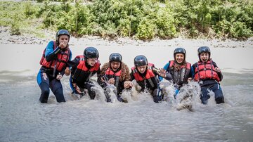 A group of six people is standing in the water and having a lot of fun. They are all wearing life vests and helmets.