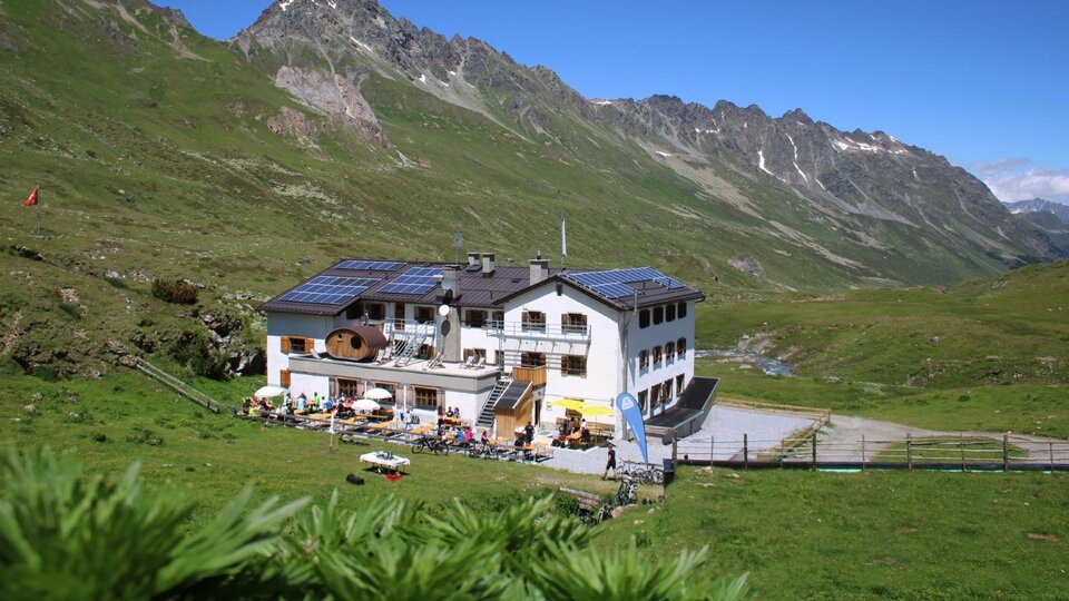 Ein charmantes Berghaus in einer grünen Landschaft. Im Hintergrund sind majestätische Berge und blauer Himmel zu sehen.