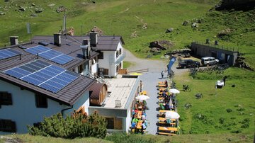 Ein rustikales Gebäude mit Solarpanelen in einer grünen Berglandschaft. Draußen stehen Tische mit Sonnenschirmen für Gäste.