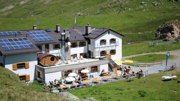 Eine Berghütte mit Terrassen und Gästen, die im Freien sitzen. Über dem Gebäude befinden sich Solarpanele und die Umgebung ist grün und hügelig.