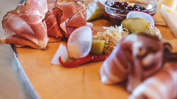 Ein schönes Holzbrett mit verschiedenen Wurst- und Käsesorten. Dazu sind Oliven und ein Dip in einer kleinen Schüssel arrangiert.