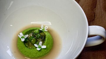 Ein elegantes Gericht in einer weißen Schüssel mit einer grünen Creme und essbaren Blumen. Der Hintergrund ist aus Holz, was dem Bild eine rustikale Note verleiht.