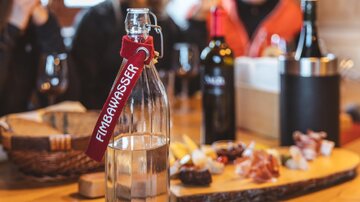 Eine Glasflasche mit Wasser steht auf einem Tisch, umgeben von verschiedenen Weinen und einer Auswahl an Snacks. Im Hintergrund sind einige Personen zu sehen, die sich unterhalten.
