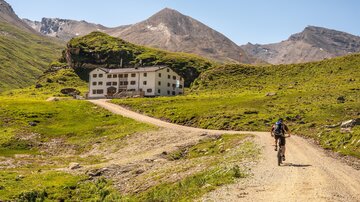 Ein Fahrradfahrer fährt auf einem schmalen Kiesweg in den Bergen. Im Hintergrund steht ein großes, altes Gebäude umgeben von grünen Wiesen und Bergen.