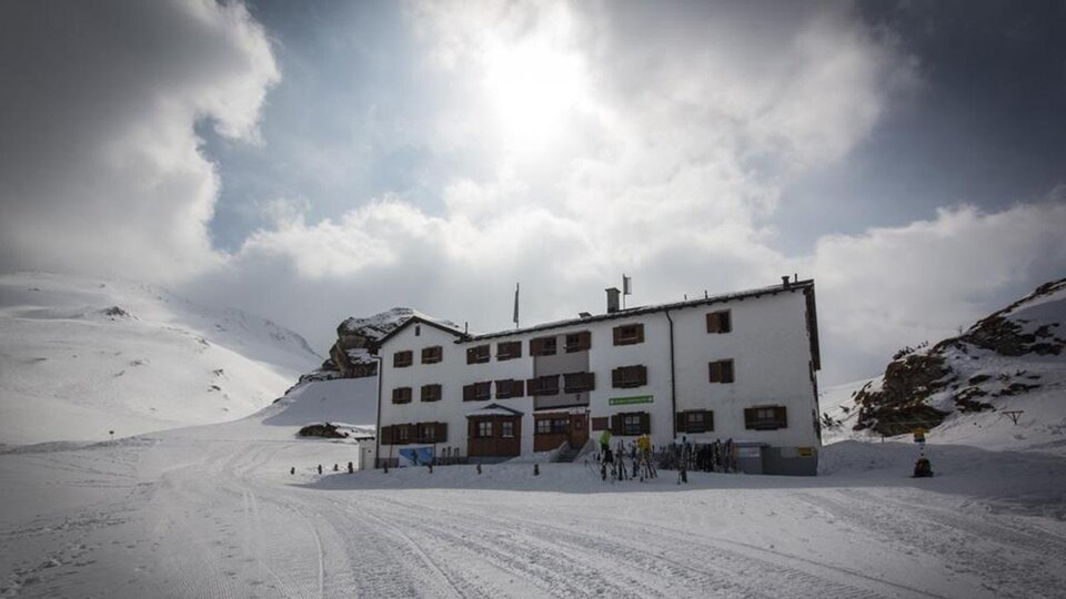 A white building in the snow, surrounded by mountains and clouds. People can be seen near the house, which radiates a cozy atmosphere.