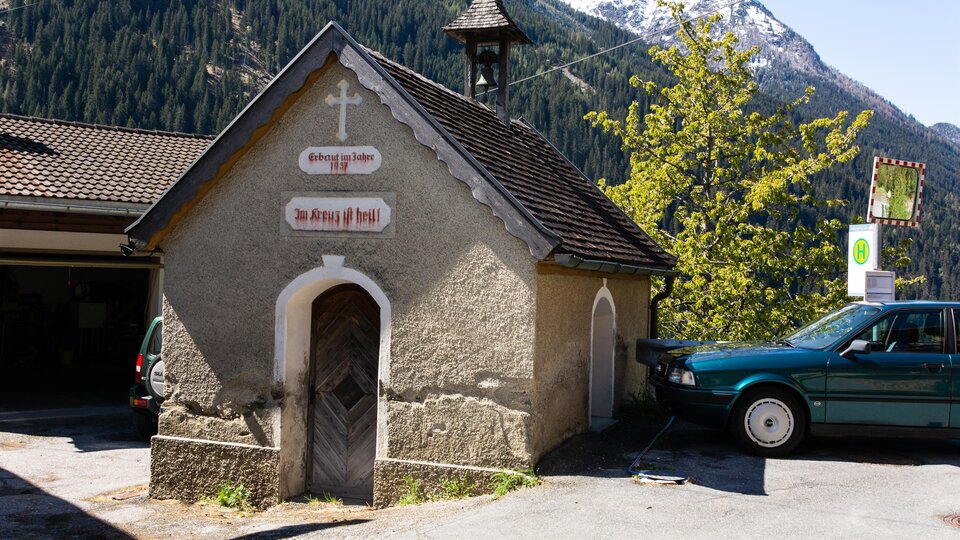A small chapel with an elegant roof shape and a cross. In the background, mountains and trees can be seen. | © Bernhard Gruber – Photography