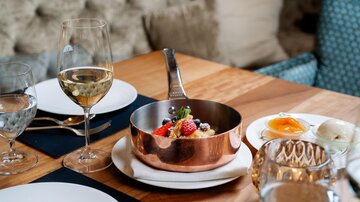 An elegantly set table with glasses, a copper-colored pot full of fresh fruits, and a white sauce. A calm atmosphere with tasteful decoration.