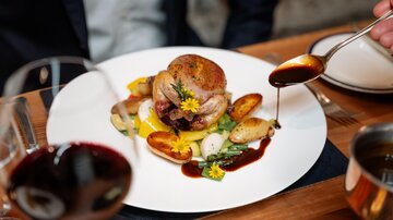 A delicious dish with roasted chicken, vegetables, and sauce on a white plate. In the background, there are wine and an elegantly set table.