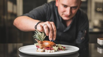 A chef decorates a carefully arranged dish on a white plate. The presentation includes fresh herbs and an appealing arrangement of ingredients.