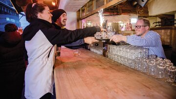 A lively bar with cheerful guests ordering drinks. A bartender serves kindly while the atmosphere is cozy.